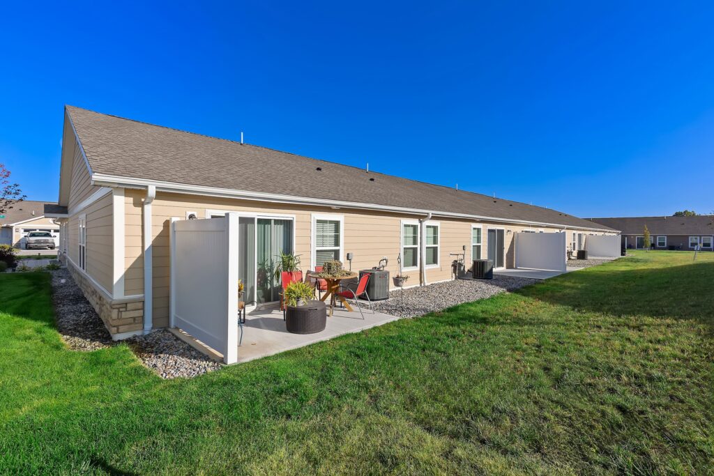 Single-story beige residential building with private patios, outdoor furniture, and landscaped grass lawn under a clear blue sky.