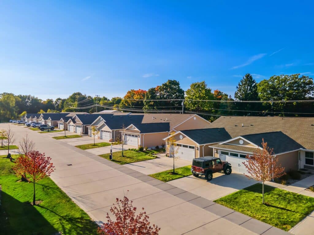 A row of single-story suburban houses with driveways and garages, lined along a clean street with green lawns and trees under a clear blue sky.