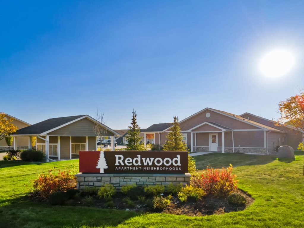 A sunny day at Redwood Apartment Neighborhoods, showing single-story residential buildings and a landscaped entrance sign on a grassy lawn.