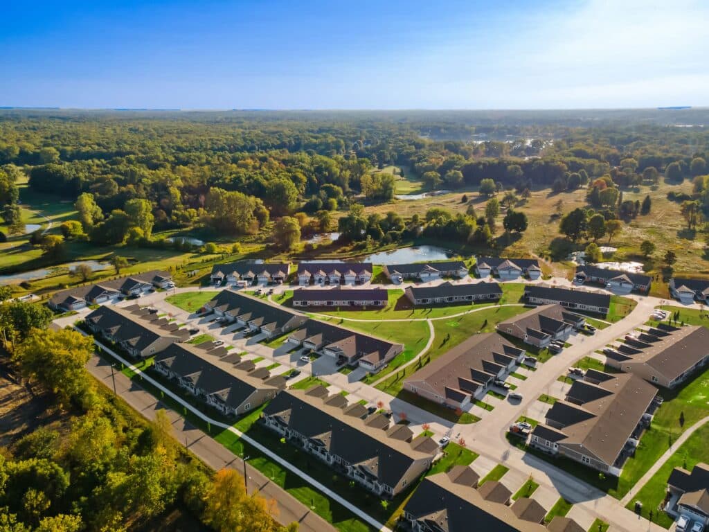 Aerial view of a suburban neighborhood with rows of houses, green lawns, curved streets, and surrounding trees under a clear sky.