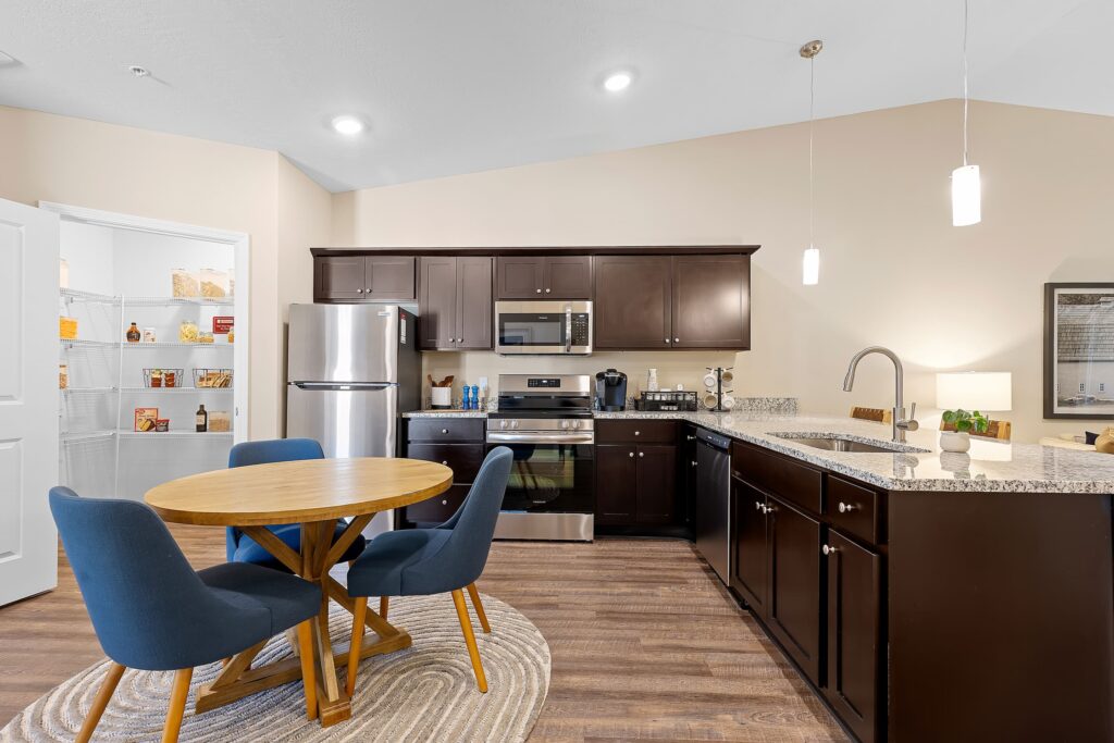 Modern kitchen with dark wood cabinets, stainless steel appliances, a round dining table with four blue chairs, and an open pantry visible on the left.