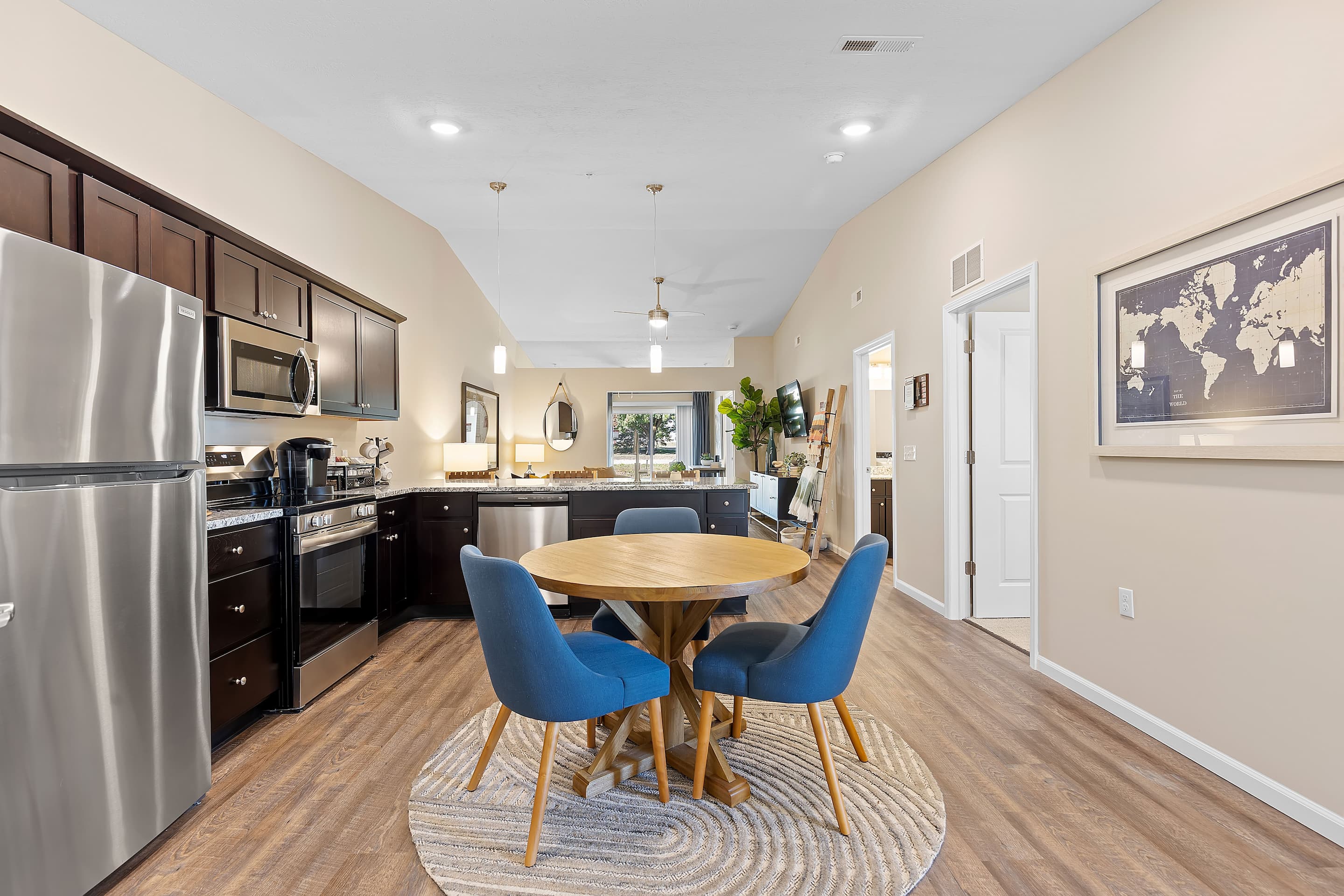 Modern kitchen and dining area with dark cabinets, stainless steel appliances, round wooden table, four blue chairs, and a world map artwork on the wall.