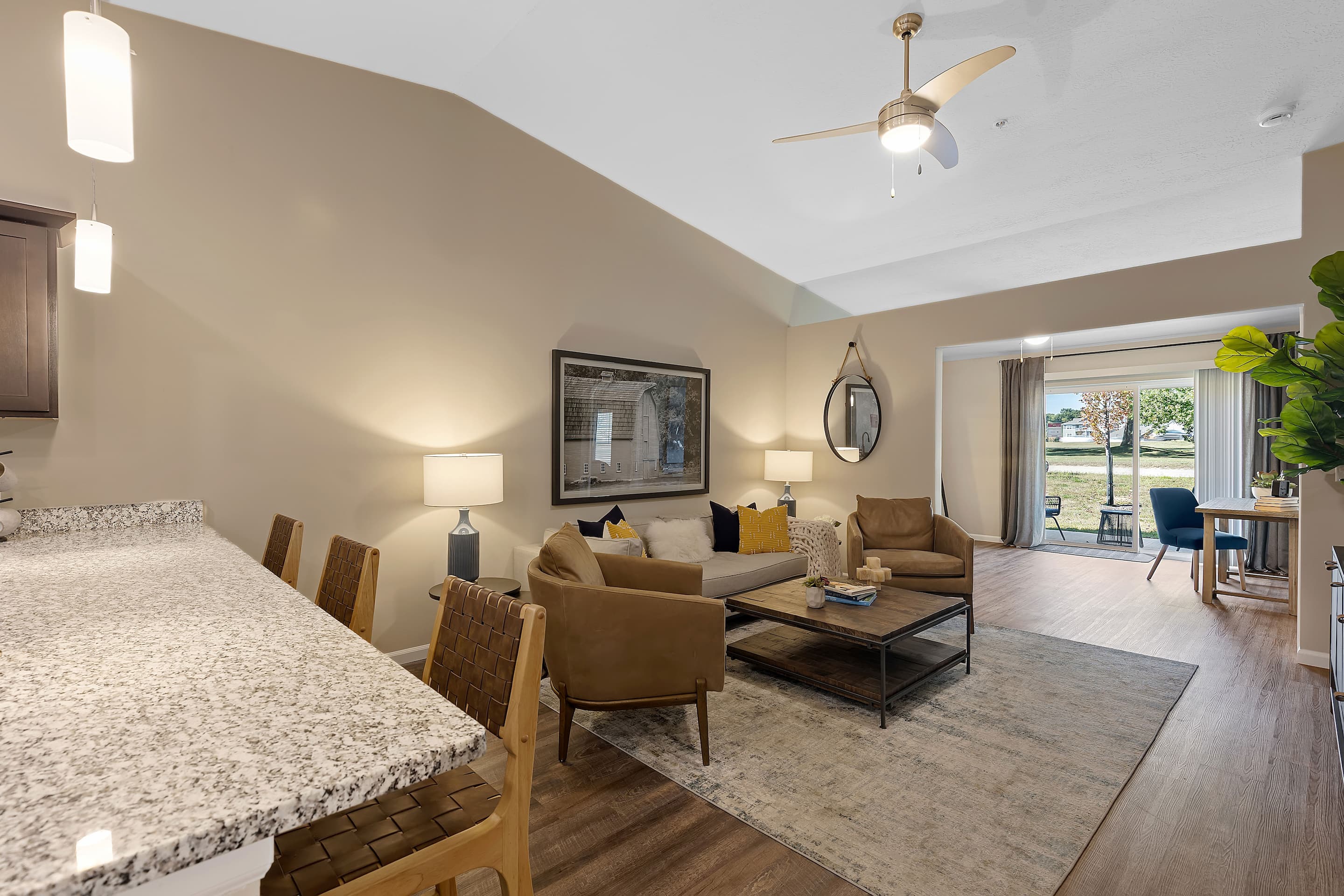 Open-concept living room with neutral walls, modern furniture, a ceiling fan, and large windows showing a view of greenery outside. Part of a kitchen counter is visible in the foreground.
