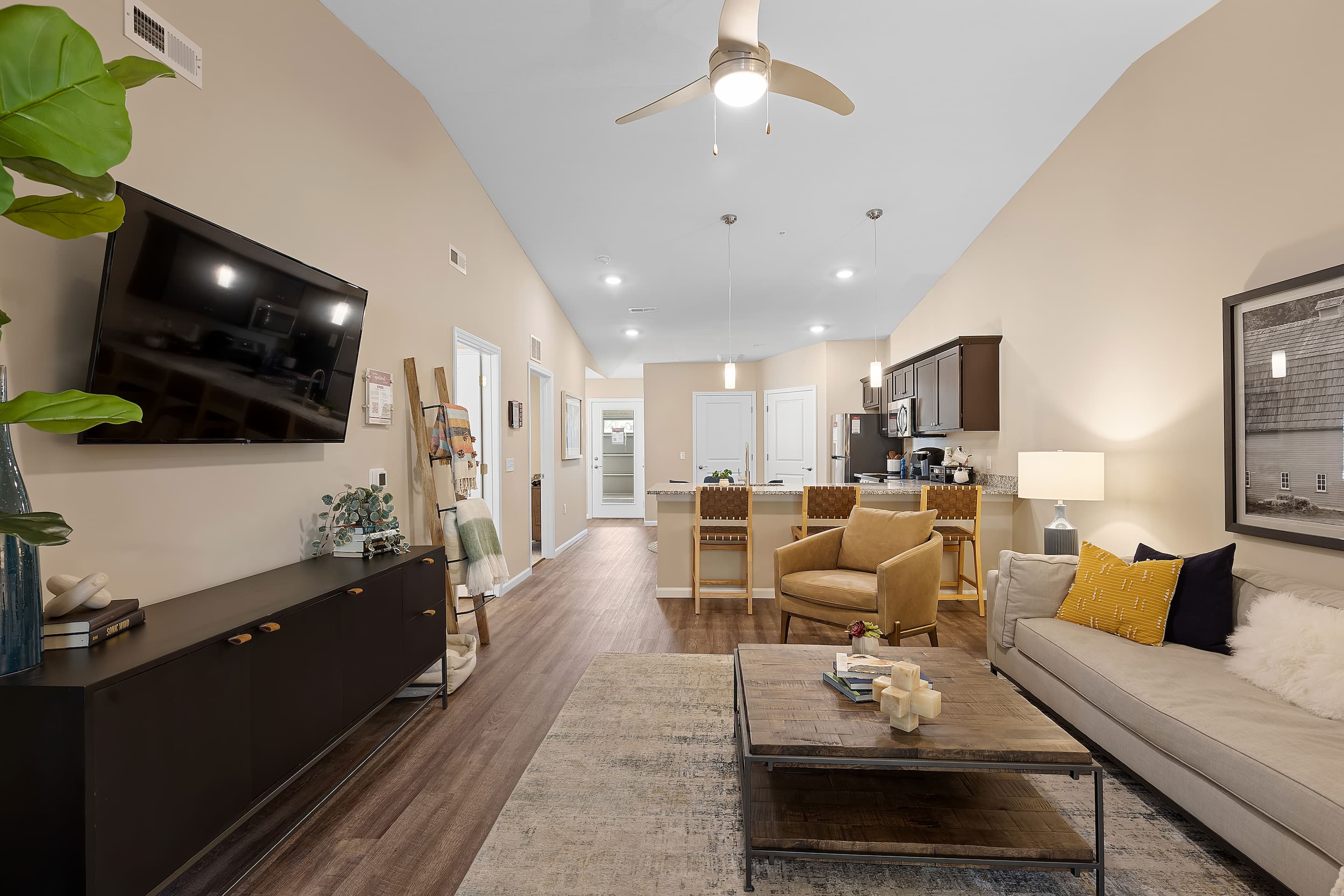 Modern open-concept living room and kitchen with neutral tones, a wall-mounted TV, sofa, armchairs, and kitchen island with bar stools under ceiling fan lighting.