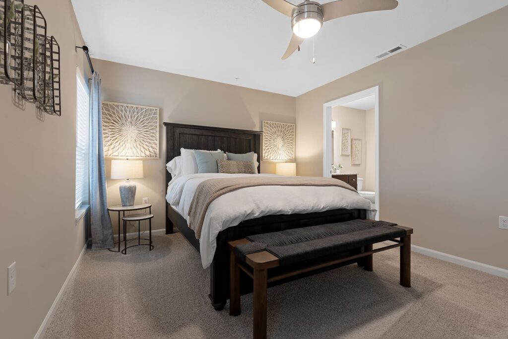 A neatly made bed with white bedding and multiple pillows sits in a carpeted bedroom, featuring beige walls, a ceiling fan, and a bench at the foot of the bed.