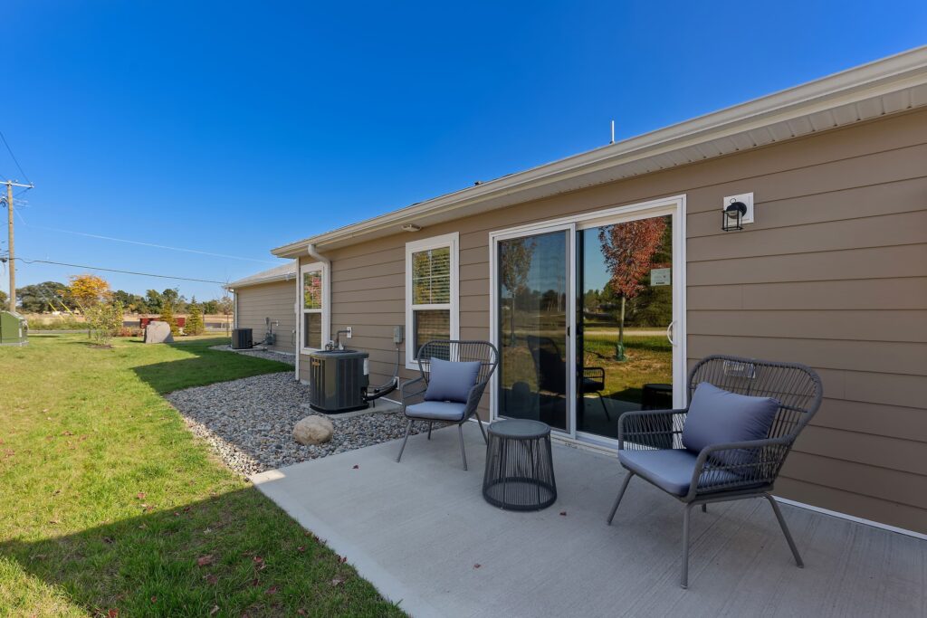 Patio area outside a tan house with two chairs, a small table, and air conditioning units by a gravel bed, facing a grassy yard and trees under a clear blue sky.