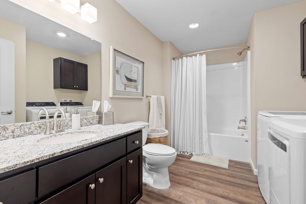 Modern bathroom with granite countertop, sink, toilet, bathtub with shower curtain, washer, dryer, wall art, and wooden floor.