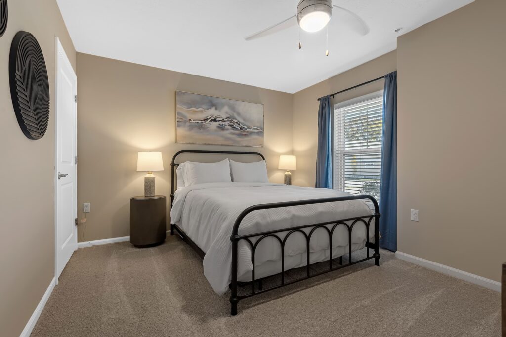 A bedroom with beige walls, a black metal bed frame, white bedding, two bedside tables with lamps, blue curtains, and a window letting in natural light.