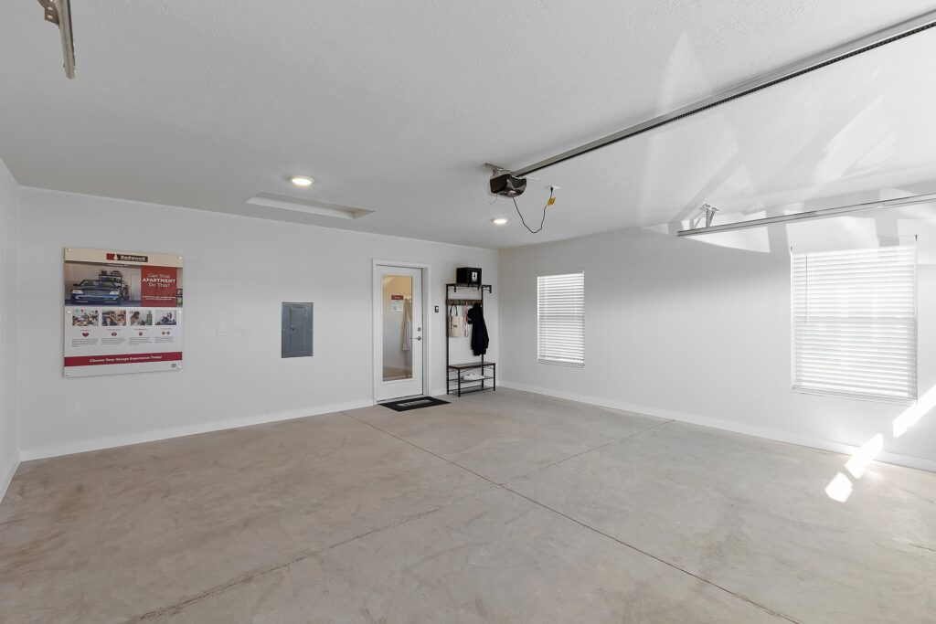 Clean, empty garage with white walls, concrete floor, two windows with blinds, a door, a wall-mounted organizer, and a bulletin board on the left wall.