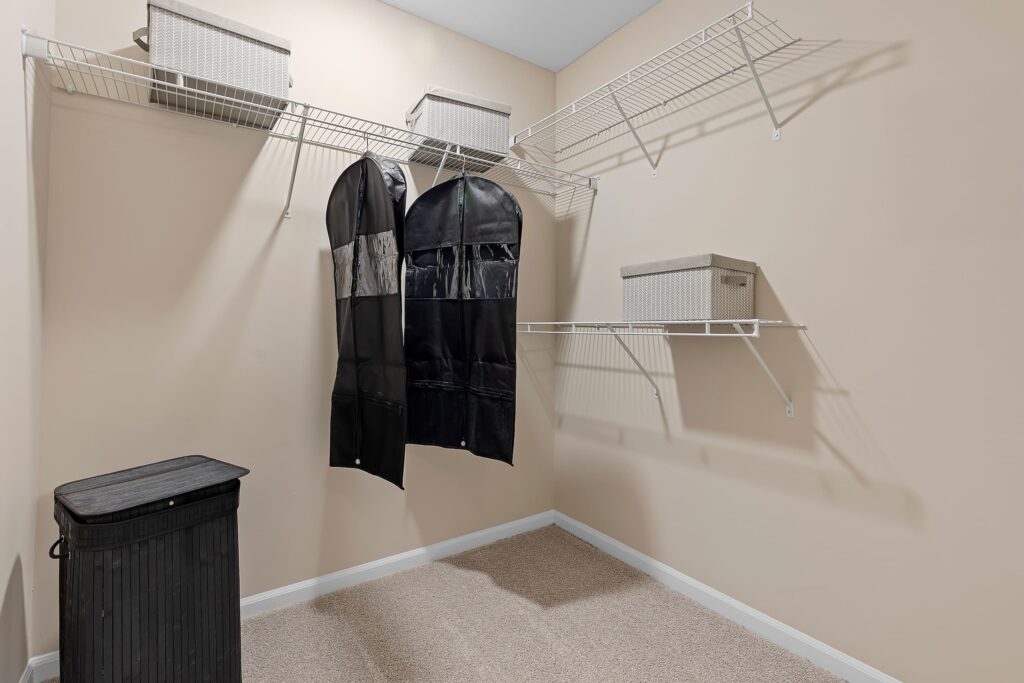 An empty walk-in closet with wire shelves, a black laundry hamper, four beige storage bins, and two garment bags hanging on the rack.