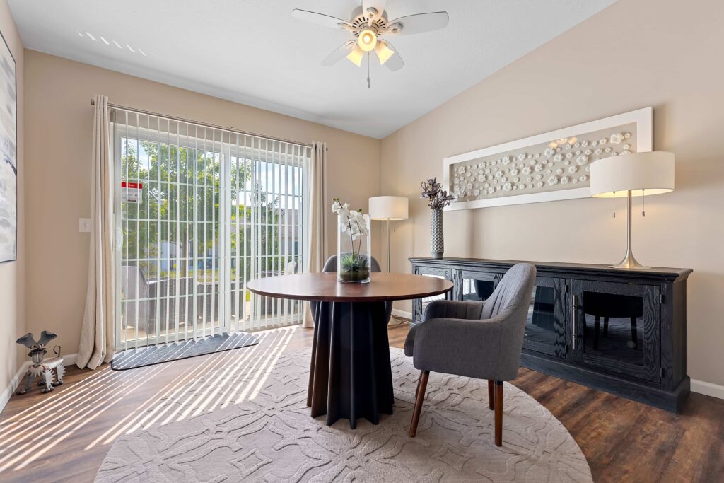 A modern home office with a round table, gray chair, black sideboard, lamps, and a large window with vertical blinds letting in natural light.