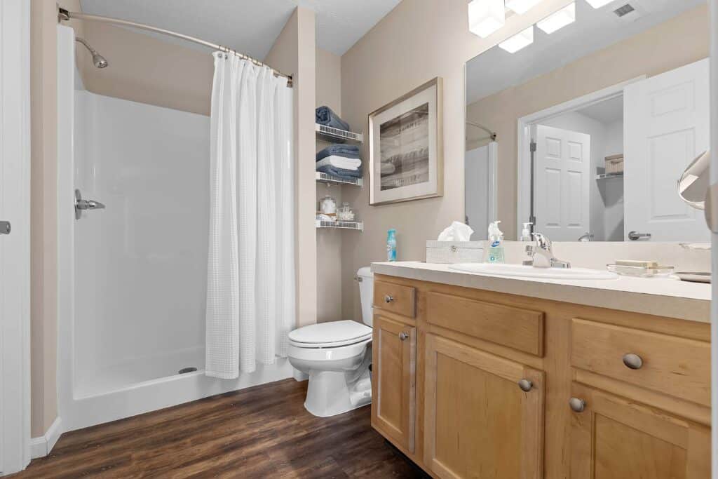 Modern bathroom with a walk-in shower, white curtain, toilet, wooden vanity, large mirror, and shelving with towels and toiletries, all set on a wood-style floor.