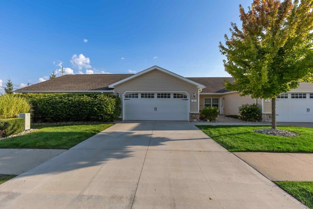 Single-story suburban house with a double garage, concrete driveway, green lawn, bushes, and a tree in front, under a clear blue sky.