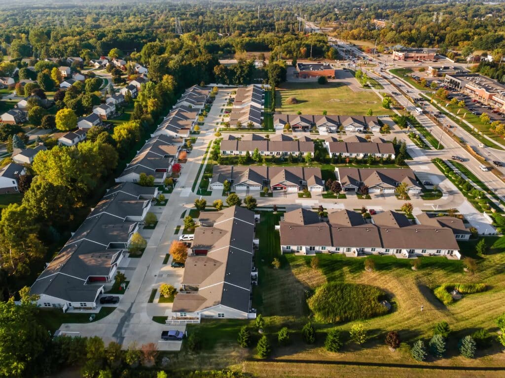 Aerial view of a suburban neighborhood with rows of houses, driveways, green lawns, trees, and nearby roads and buildings.