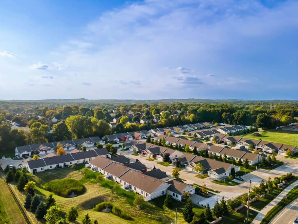 Aerial view of a suburban neighborhood with rows of single-story houses, trees, and roads surrounded by greenery under a clear blue sky.