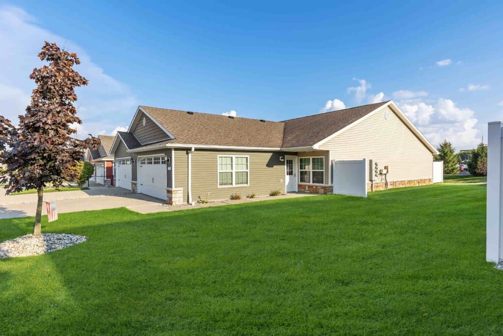 Single-story suburban house with an attached two-car garage, tan siding, a small front porch, and a well-kept lawn under a clear blue sky.
