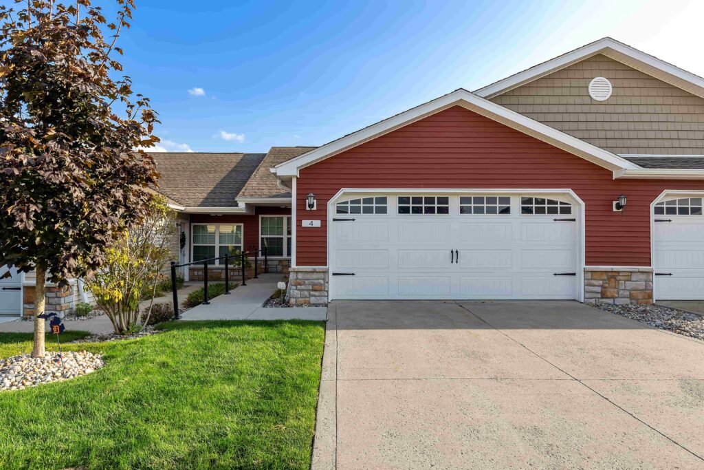 Single-story house with red siding, white garage doors, a concrete driveway, and a small front lawn with landscaping and a tree.