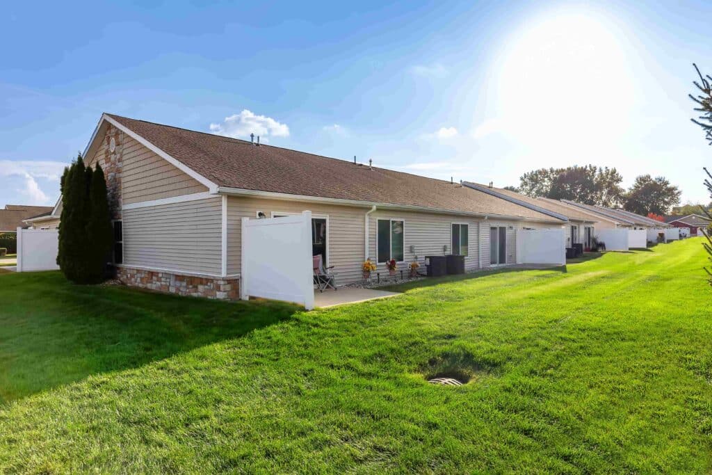 Row of single-story townhouses with beige siding, private patios, and white privacy panels, set on a well-maintained green lawn under a sunny sky.