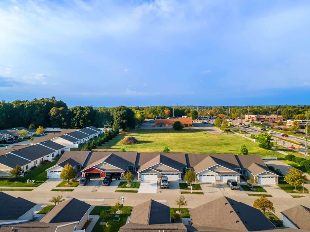 Aerial view of a suburban neighborhood with rows of houses, driveways, green lawns, and a grassy field in the background under a partly cloudy sky.