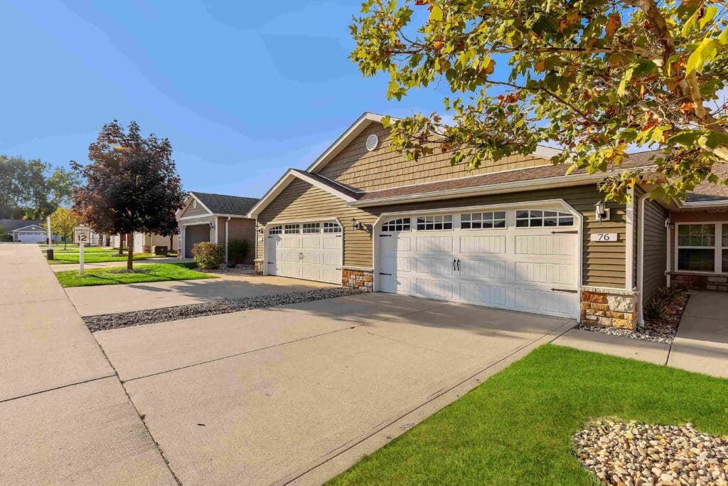 A row of suburban houses with attached garages, manicured lawns, and a tree-lined street under a clear blue sky.