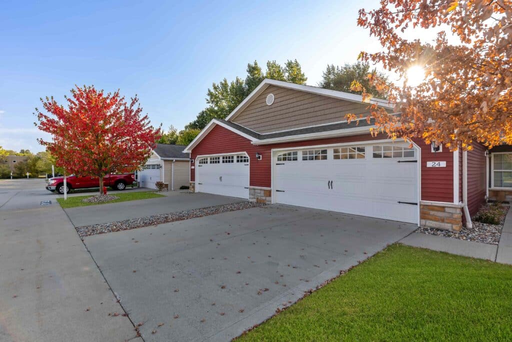 A red duplex with white garage doors and a driveway, bordered by green grass and trees with autumn foliage, under a clear sky.