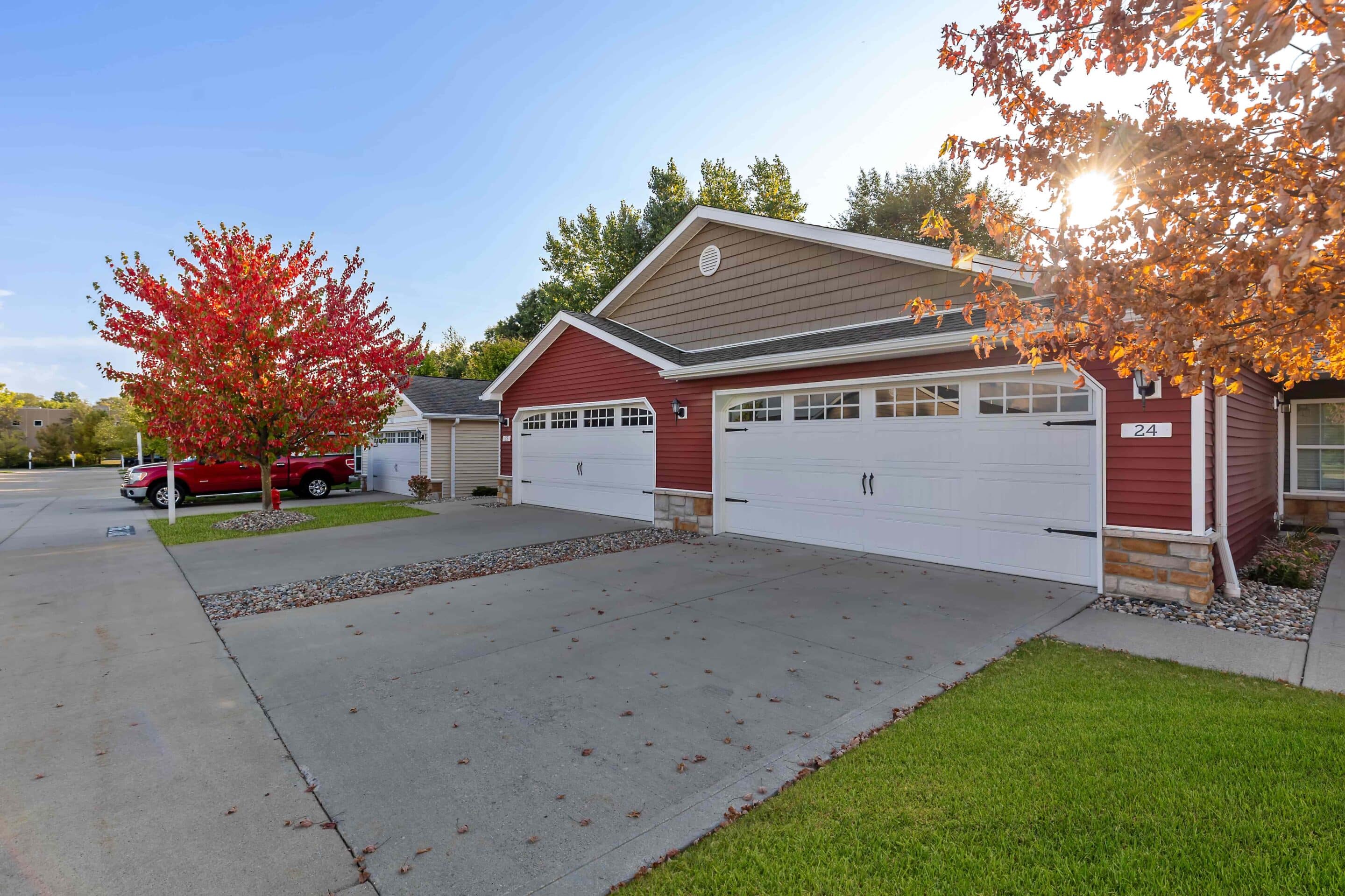 A red duplex with white garage doors and a driveway, bordered by green grass and trees with autumn foliage, under a clear sky.