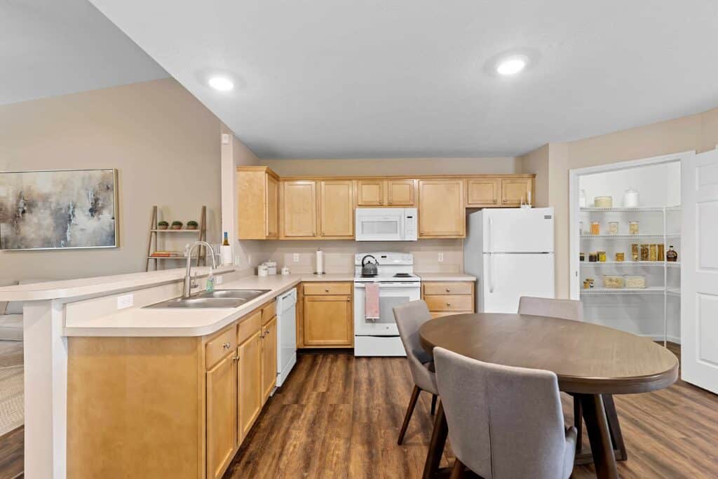 A kitchen with light wood cabinets, white appliances, a round dining table with four gray chairs, and an open pantry with shelves.