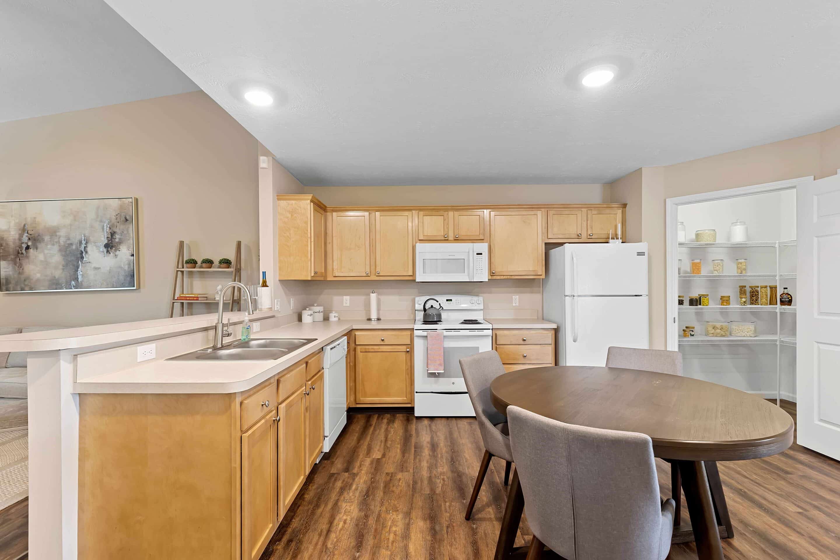 A kitchen with light wood cabinets, white appliances, a round dining table with four gray chairs, and an open pantry with shelves.