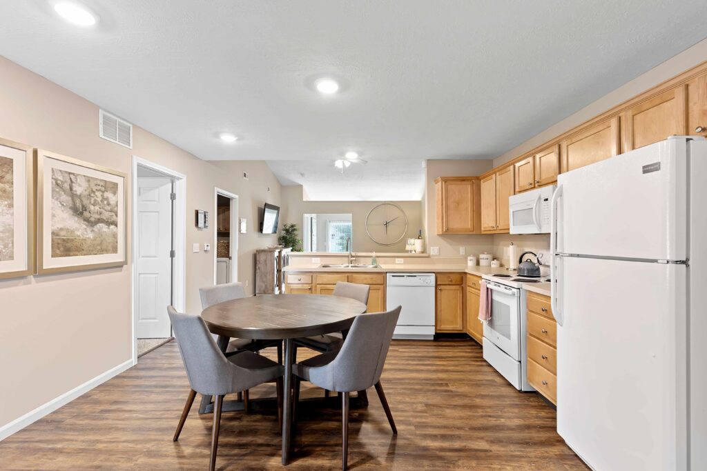 Modern kitchen and dining area with wooden cabinets, white appliances, a round dining table with four gray chairs, wood-style flooring, and wall art.