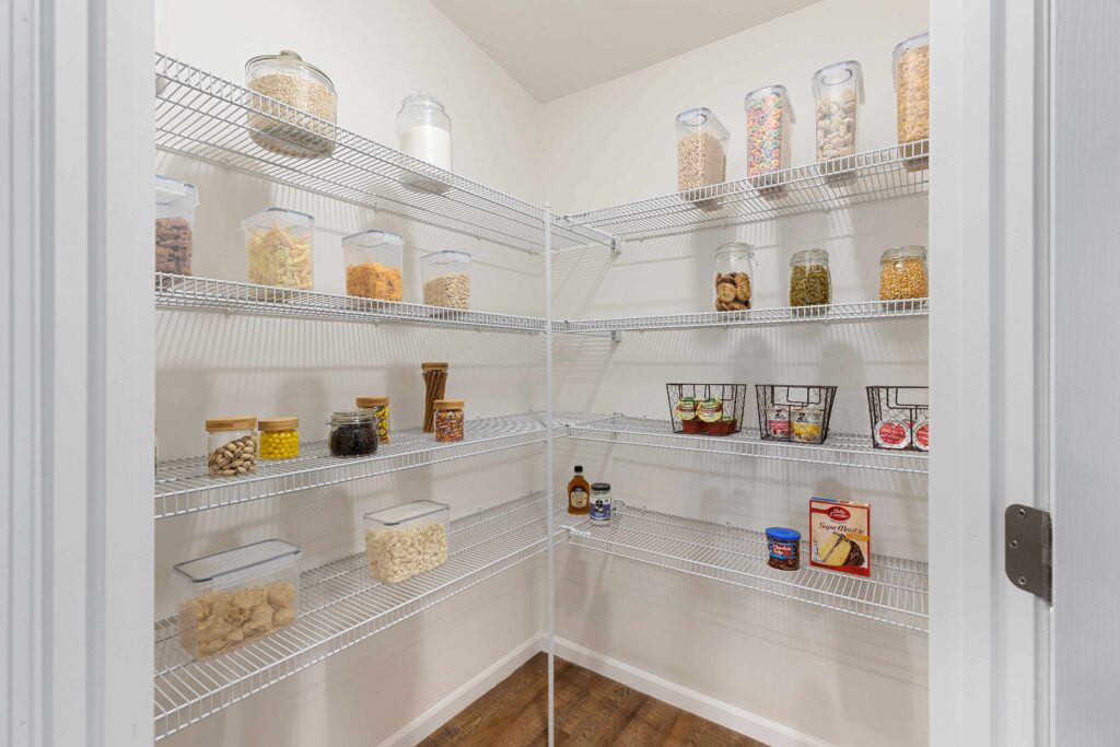 A neatly organized pantry with wire shelves holding assorted jars, containers, and baskets filled with food items against white walls and a wooden floor.