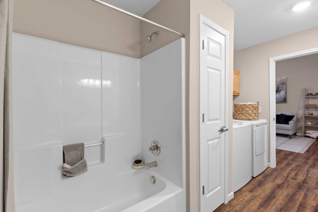 A bathroom with a white bathtub and shower, adjacent to a laundry area with a washer, dryer, and wicker basket, leading to a living space.