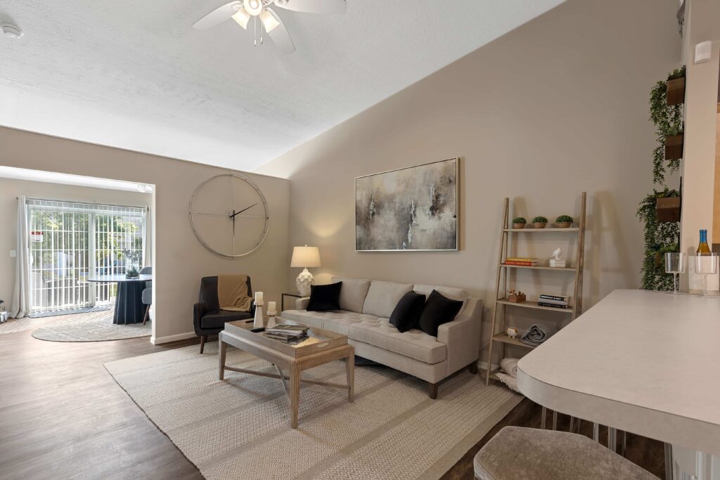 A modern living room with a beige sofa, black and beige cushions, a coffee table, abstract wall art, a large wall clock, and a view into a sunroom with sliding glass doors.