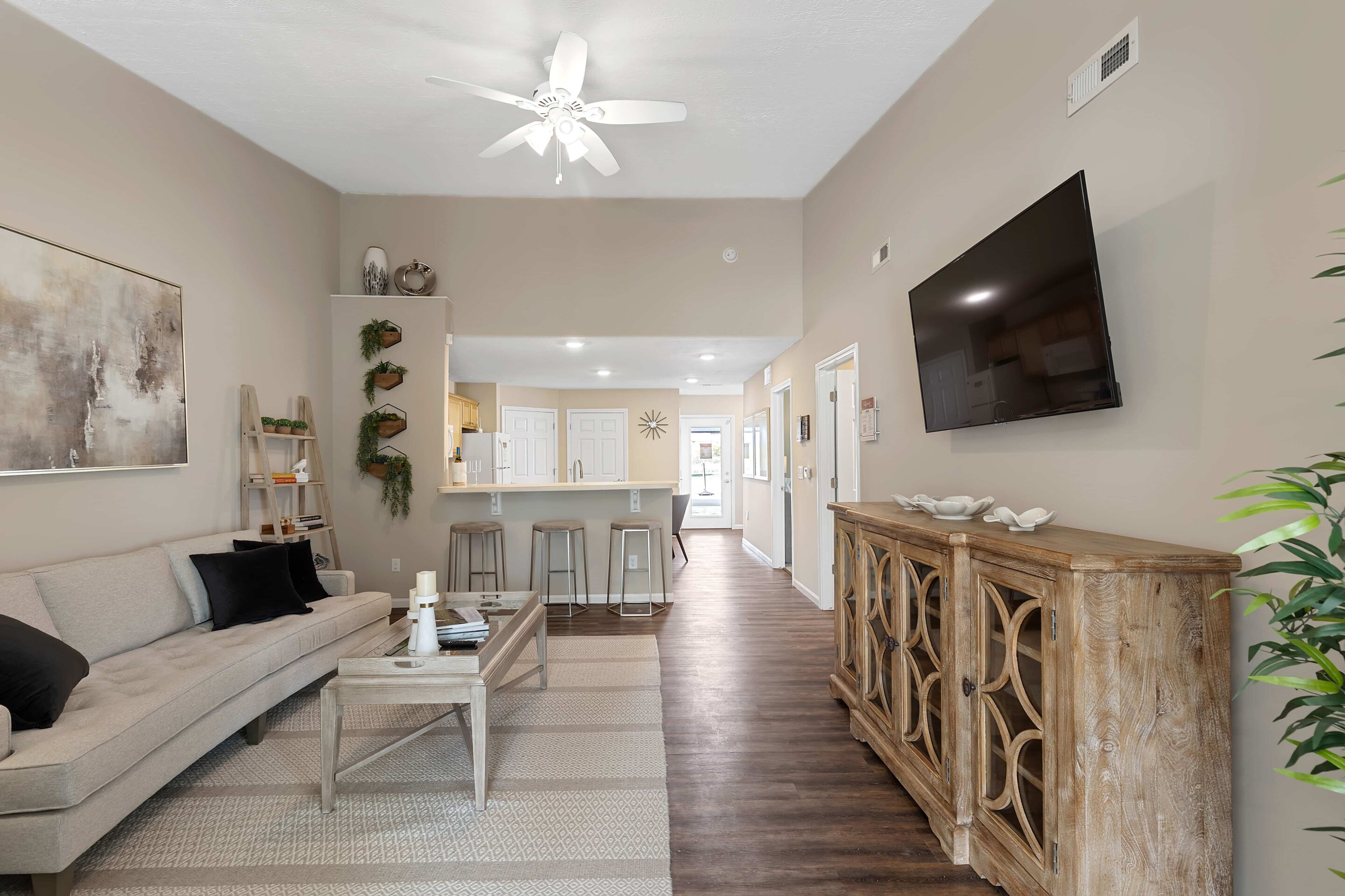 Modern living room with neutral decor, a beige sofa, wall-mounted TV, wooden sideboard, and open view into a kitchen with bar stools and pendant lighting.