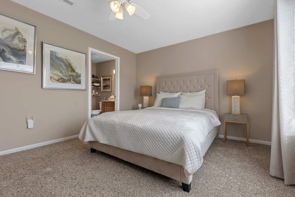 A neatly made bed with white bedding in a carpeted bedroom, featuring bedside tables with lamps, artwork on the walls, and an open door to a bathroom.