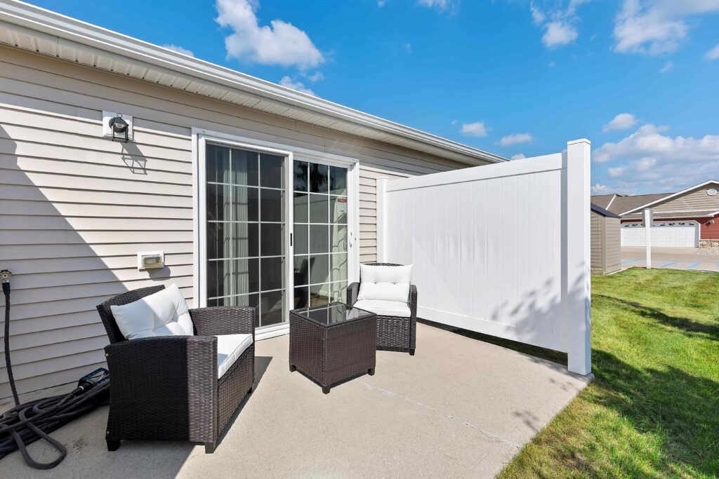 Small patio area with two cushioned chairs, a glass-top table, and a white privacy fence next to a sliding glass door of a beige house under a blue sky.