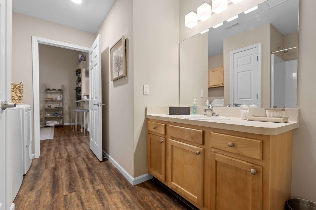 A bathroom with light wood cabinets, a sink, mirror, and overhead lights. The room features wood flooring and leads to a laundry area with shelves and a washing machine.