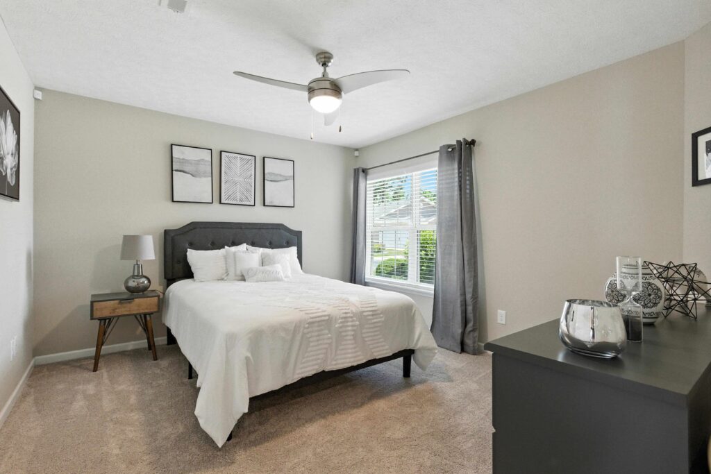 A neatly made bed with white linens sits beside a nightstand in a carpeted bedroom with neutral walls, a ceiling fan, dresser, and window with gray curtains.