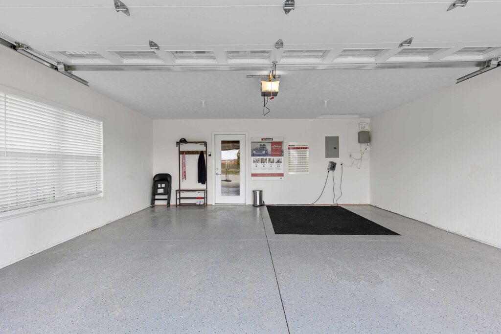 Clean, empty garage with gray floor, white walls, large window, black mat, and a wall-mounted coat rack near a door with a poster. Ceiling lights and garage door opener are visible.
