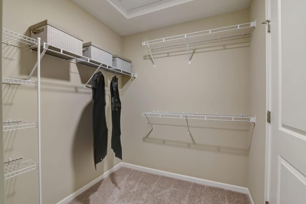 Empty walk-in closet with beige walls, carpeted floor, wire shelves, two hanging garments, and three storage bins placed on the top shelf.