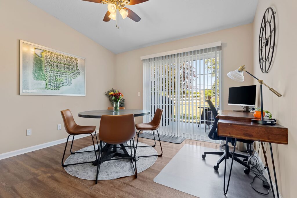 A modern home office and dining area with a round table and four chairs, a desk with a computer, wall art, and sliding glass doors with vertical blinds letting in natural light.
