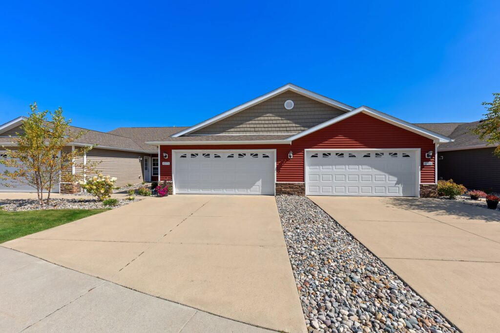 Two attached suburban houses with red and tan siding, each featuring a double garage, concrete driveways, and a rock landscaping strip between the drives. Clear blue sky overhead.
