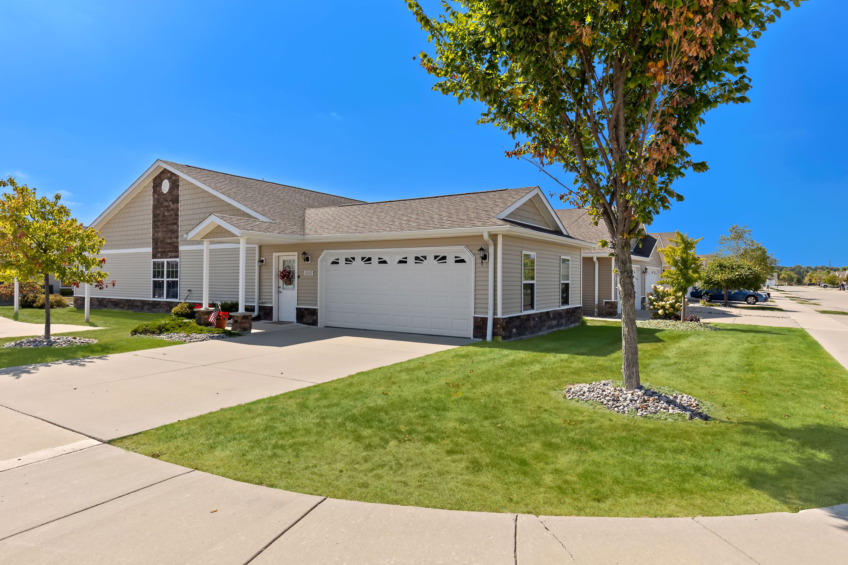 Single-story suburban house with a two-car garage, well-maintained lawn, and tree in front, on a sunny day.