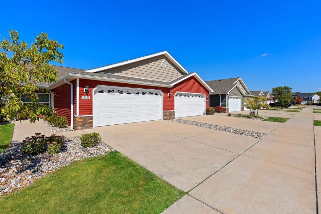 Row of suburban single-story houses with double garages, red and beige exteriors, landscaped lawns, and clear blue sky.