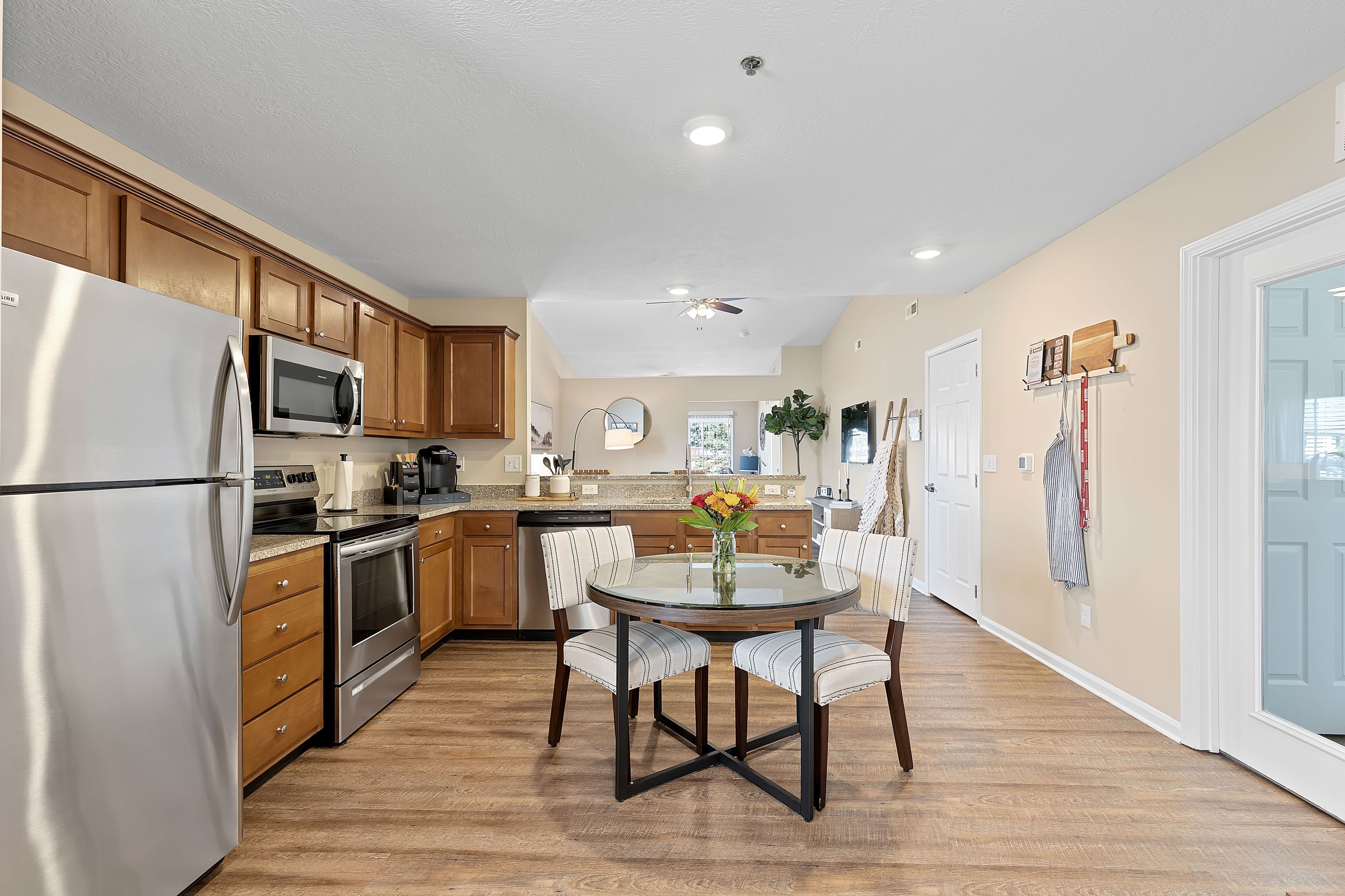Modern kitchen and dining area with wooden cabinets, stainless steel appliances, a round glass table with four chairs, and a vase of flowers, all on wood flooring.