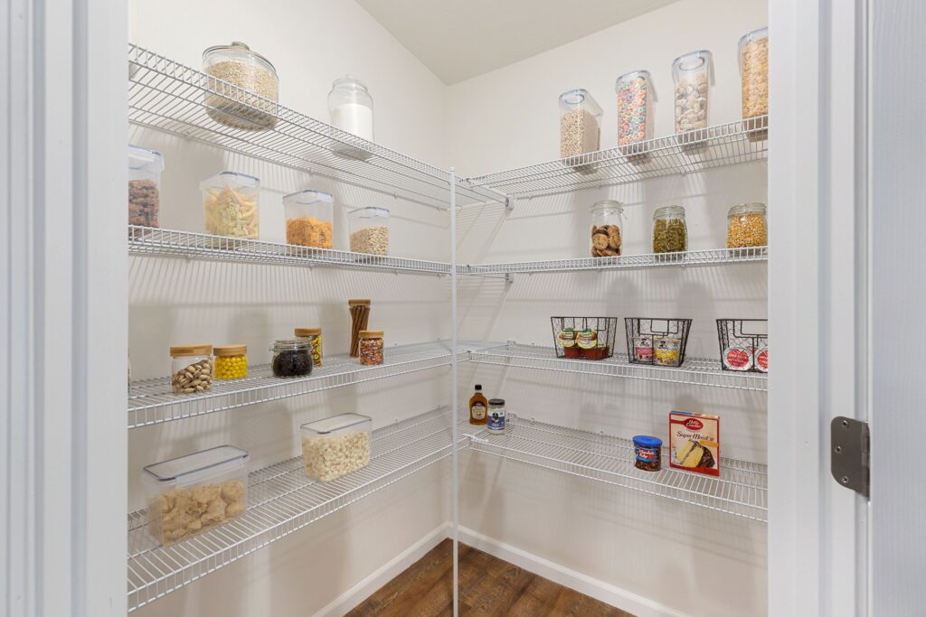 A neatly organized pantry with wire shelves holding clear jars of dry goods, baskets of canned items, and a few bottled condiments on wooden flooring.