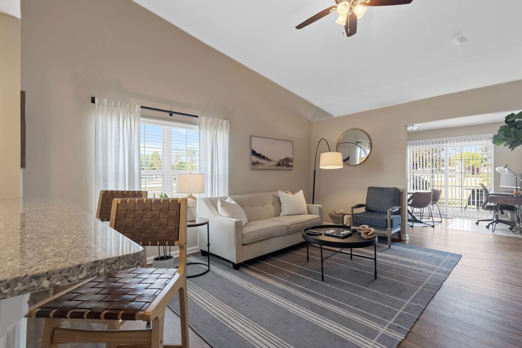 Modern living room with neutral decor, a white sofa, armchair, round coffee table, striped rug, and large windows with sheer curtains letting in natural light.