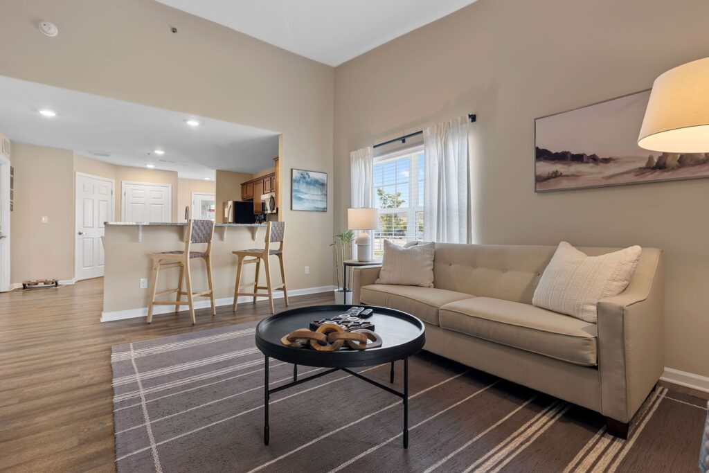 A modern living room with a beige sofa, round black coffee table, floor lamp, and two barstools at a kitchen counter; large window with white curtains lets in natural light.