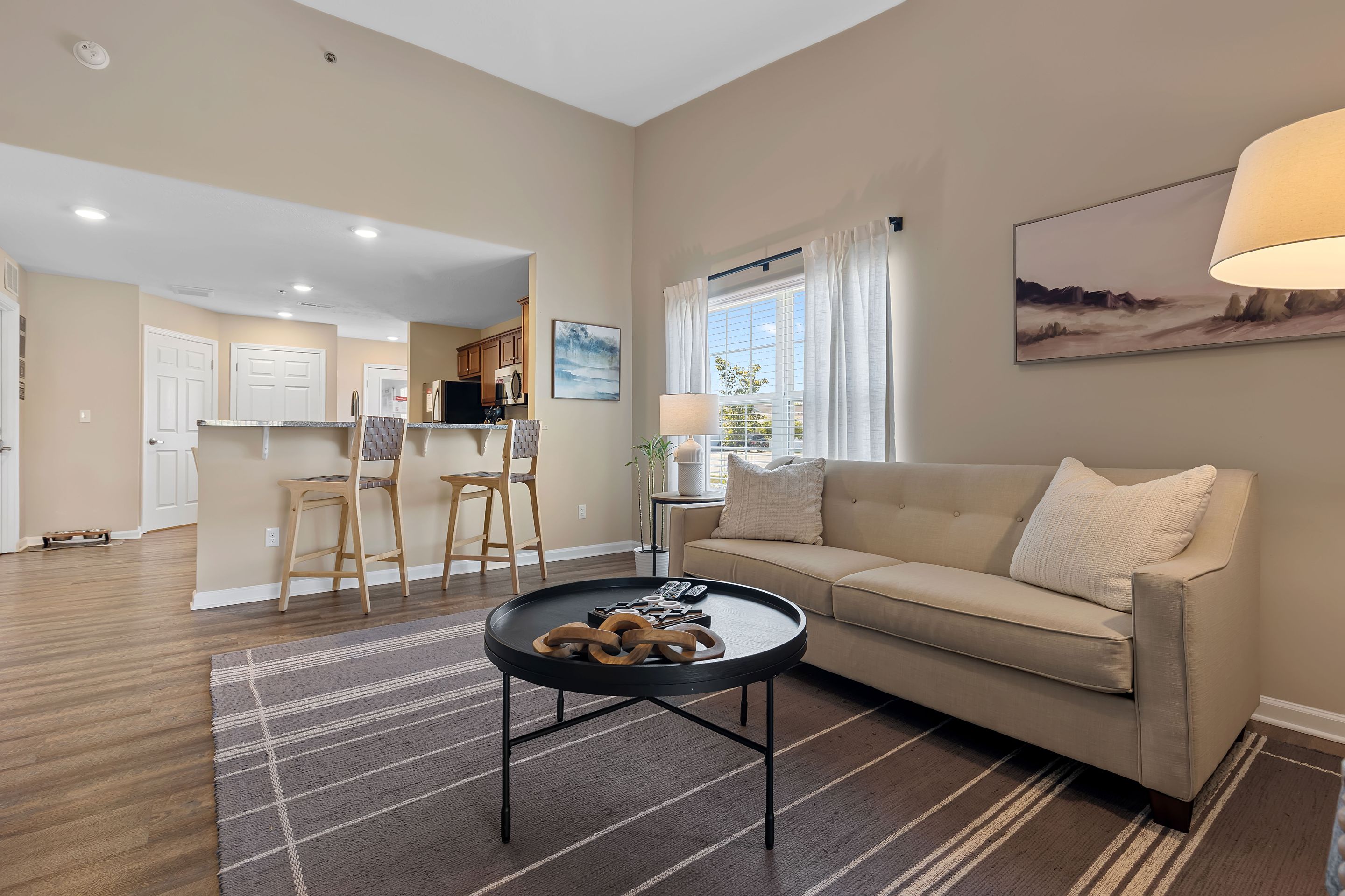 A modern living room with a beige sofa, round black coffee table, floor lamp, and two barstools at a kitchen counter; large window with white curtains lets in natural light.