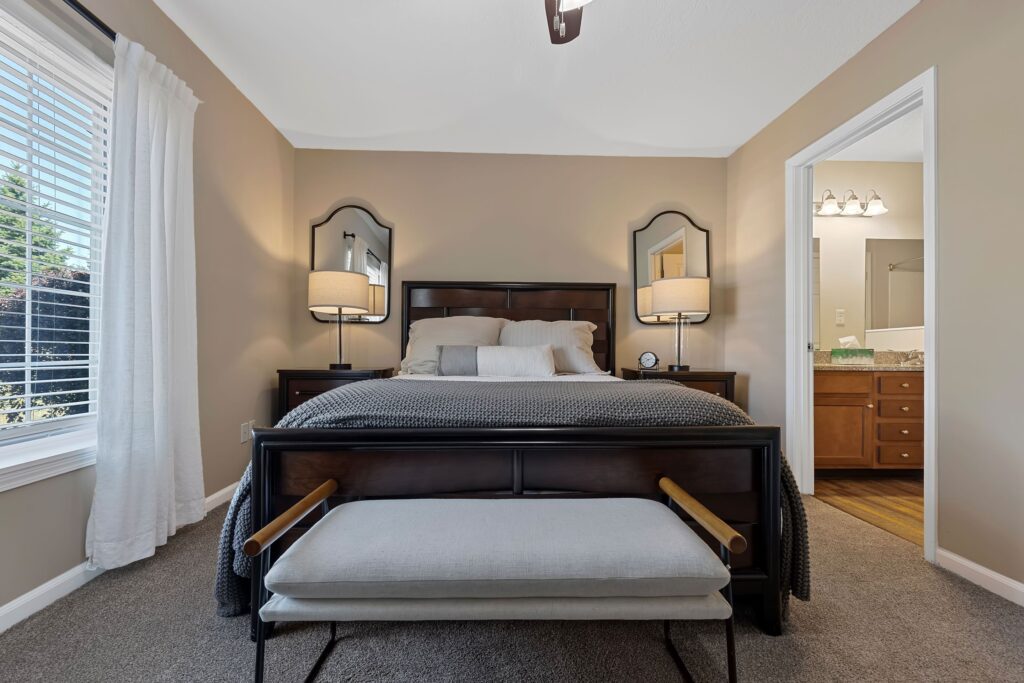 A neatly made bed with pillows and a gray throw blanket is centered in a bedroom, with two nightstands, lamps, mirrors, and a bench at the foot of the bed. An ensuite bathroom is visible.