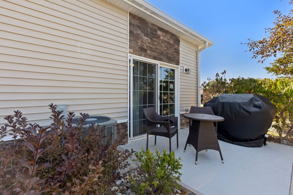 Small patio with two wicker chairs, a round table, a covered grill, an air conditioning unit, and surrounding plants, adjacent to a house with sliding glass doors.